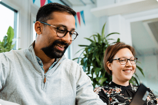 Two people sitting indoors, smiling while looking at something off-camera. They are wearing glasses and casual clothing, with plants and decorations in the background.